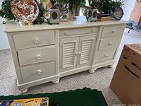 Front view of a white distressed dresser with seven drawers and two central cabinet doors with louvered panels. Decorative plates and decor on top.