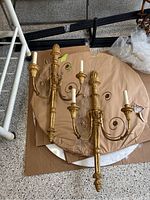 Two gold-painted vintage wall sconces with three curved arms and candle-style sockets lying on cardboard.