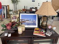 Overview of lot items including lampshade, framed photo, wine glasses, books, and decorative vase with faux greenery and feathers on a dark wooden cabinet.