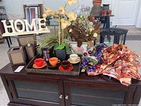 Overview of the lot showing tablecloths, napkins, napkin rings, four colored cups and saucers on a serving tray, two lantern candle holders, wooden HOME letters, and potted faux flower arrangements