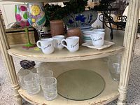 Two shelves displaying assorted home décor items including distinctive colorful wine glass with polka dots, white espresso cups, clear tumblers, brown candles, grape-themed vase, dark red wine glass holders, greenery garland, and decorative storage boxes.