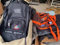 Photo showing pile of bags with a black backpack embroidered with '2006 Greenway American Century Championship' and 'NBC Sports' logos on top.