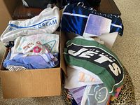 Two boxes filled with folded pillows and towels showing different colors and patterns, prominently featuring New York Jets and Pikachu themed items.