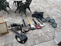 Wide photo showing 4 guitar game controllers and a 5-piece drum set controller laid on stone tiles outside