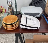 Lot contents on brown table showing George Foreman grills, Hampton milkshake maker, stacked yellow brown ceramic dishes with chips, and black paper towel holder.