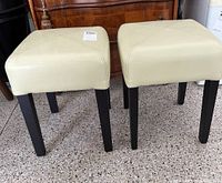 Photo showing two cream leather top stools with square seats and black legs on a tiled floor.