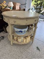 Front view showing round table with two drawers and two shelves holding decorative items and candles. Ornate carved edge detail visible around top surface.