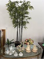 Photo shows tall artificial bamboo plant, white ceramic tea service items, floral arrangement on glass table.