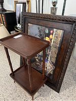 Two-tiered wooden table with spindle legs placed in front of a framed wall art leaning against wall.