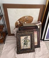 Ten pieces of wall art stacked and leaning, showing a large leaf painting in the back and ornate framed paintings in front