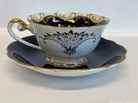 Tea cup showing front side with gold and black floral and scroll designs on white porcelain and scalloped gold trimmed rim and handle.