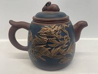 Front view of brown and dark blue ceramic teapot with detailed raised dragon design on the body, curved handle and spout, lid with sculpted figure.