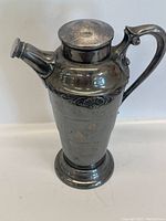 Front angle of tall silver plated cocktail shaker with decorative band and hinged lid closed