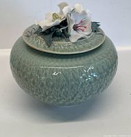 Front view of the jar with the lid on, showing textured light greenish-blue porcelain body and white porcelain flowers on lid
