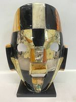 Front view of vintage mask with semi precious stones inlaid in a checkered pattern, mounted on a black base.