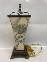 Full view of the lamp showing the trapezoidal fabric shade and dark metal base with finial, including power cord and switch.