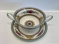 Bone china cream bowl with two handles, floral and gold ornamental design interior and exterior, white background with green rim