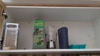 Shelving cabinet with various kitchen tools including metal box grater, Top Chop chopper in packaging, and clear plastic pitchers.
