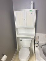 Full view of white laminated wood cupboard installed over a toilet, showing top cabinet with two paneled doors, open middle shelf, and white finish.