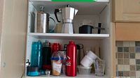Wide view of two shelves in a cabinet showing espresso maker, milk frother, multiple water bottles, measuring cups, and other small kitchen items