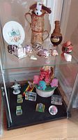 Glass cabinet shelf with six glass inside etchings, cups, decorated shells, a jar, plate and figurines on ceramic stand