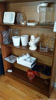 Wooden shelving unit with multiple glass and ceramic items: pitcher, tea cups with saucers, vases, plant pots, angel figurine, glass skull, coasters, and small boxes.