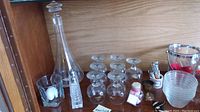 Photo showing glass decanter with stopper, six brandy glasses, and salt and pepper shakers on a wooden shelf