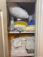 View of linen closet shelves filled with folded towels, pillowcases, comforters, and pillows mostly in white, yellow, and blue colors.