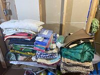 Overview of assorted folded linens including fleece blankets, dish towels, table cloths, and seat cushions stacked on a small table/bench.