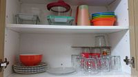 Photo of middle shelf showing stacked white plates with one red bowl, clear glass drinking glasses and snack plates.