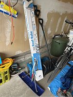 Four snow removal tools: three snow shovels leaning against the wall and one boxed Poly Roof Rake.