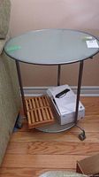 Full view of metal frame table with round glass top and lower glass shelf showing some items placed on lower shelf, positioned adjacent to sofa on wood floor.