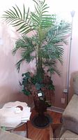 Full view of tall faux tree with green leafy branches and decorative planter base on floor next to furniture