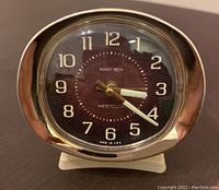 Front view of Baby Ben alarm clock showing round brown dial with white numbers and luminous hands, metal trim, and rectangular base.