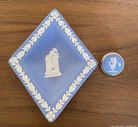 Photo of the diamond-shaped blue Wedgwood Jasperware tray with white relief decoration and the matching round brooch or pendant on a wooden surface.