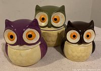 Three ceramic owl storage jars in green, purple, and brown colors photographed together on a plain background.