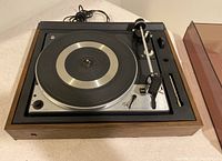 Full top view of the Dual 1225 turntable showing wood veneer base, metal controls, platter, tonearm assembled with stylus, and attached power cord.