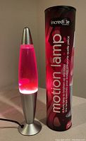 Silver base lava lamp on, pink wax clumps floating in clear liquid, next to original branded packaging tube in maroon color with 'motion lamp' text.