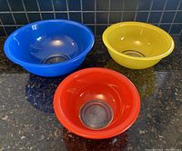 Three vintage Pyrex glass mixing bowls in red, yellow, and blue colors with clear bottoms shown on countertop.