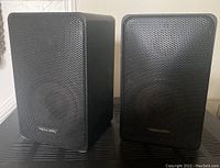 Pair of Realistic Minimus-7 speakers viewed head-on showing front metal mesh grilles with Realistic badge on one grille.