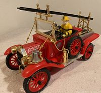 Side angle showing red vintage fire truck toy with brass colored fittings, firefighter figure, ladders, hose reel, and spare tire with New Bright branded tires.