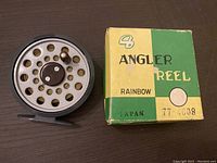 Front side view of the Rainbow #72 vintage fly fishing reel next to its original box showing perforated spool and metal handle.