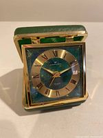 Front view of vintage Hamilton travel alarm clock showing green square folding case, gold trim, and round dial with Roman numerals.