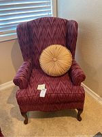 Wing-back armchair with beige round pleated pillow, upholstery showing zigzag pattern in reddish-purple fabric, wooden cabriole legs, positioned next to a window with blinds.