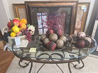 Full view of three ornate pedestal vases on a glass table, each filled with artificial fruits including grapes, apples, and lemons, with framed artworks in the background.