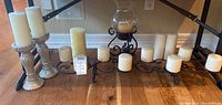 Full view of assorted candle holders and pillar candles arranged on floor. Includes clear glass textured holders, black metal scroll holders, and multiple ivory candles.