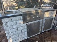 Full front view of the outdoor kitchen unit showing stone veneer cabinets, sink on the left, grill in center and fridge on right