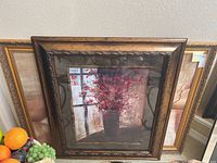 Framed still life artwork showing red flowers in a vase by a window with brown ornate framing