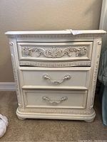 Photo front view of single cream white nightstand showing three drawers with decorative ornate carving and one drawer pulled open showing wooden interior.
