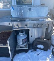 Front view of stainless steel propane grill with lid closed, snow on top, propane tank inside the cabinet with door open, grill cover and cooking accessory tray visible